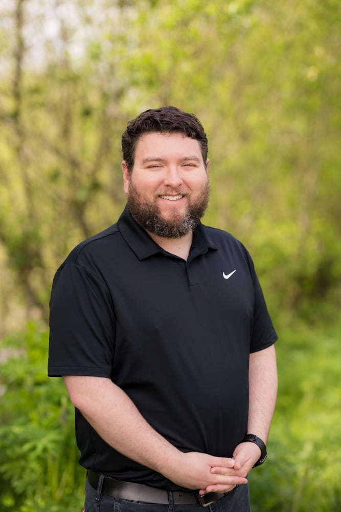 Portrait of a bearded man in a black polo shirt, standing outdoors in a green, wooded area with a friendly smile.