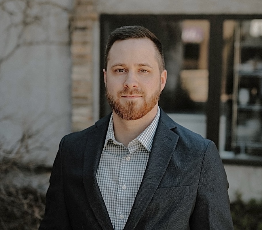 Head-and-shoulders portrait of a man in a navy blazer and checkered shirt standing outdoors.