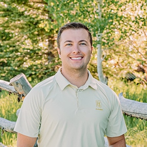 Smiling man wearing a light polo shirt stands outdoors near a wooden fence with trees in the background.